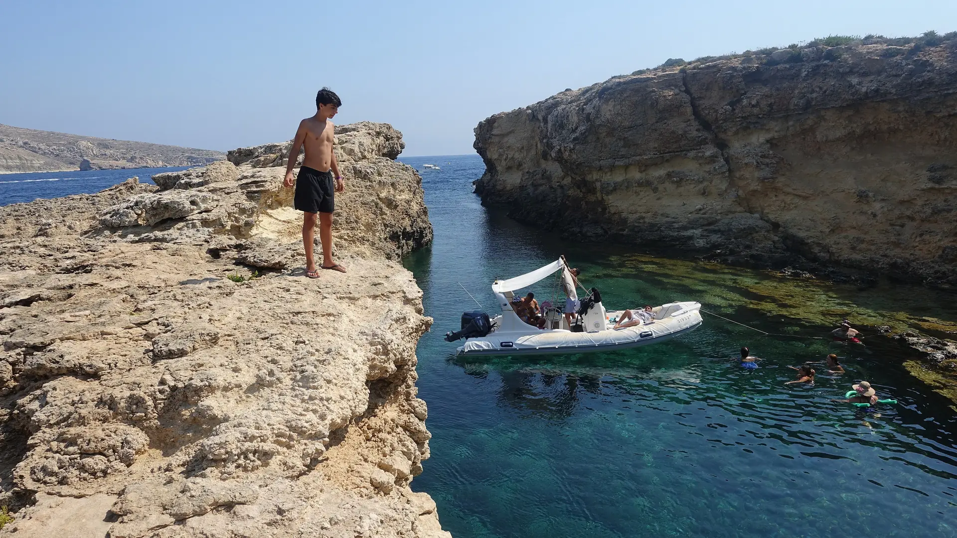 A white inflatable boat with a canopy anchored in a narrow rocky inlet on Comino, with swimmers in clear water beside it